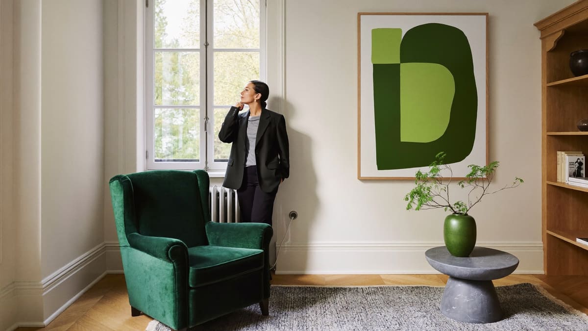 Heller Altbauraum mit grünem Samtsessel und grünem Kunstwerk an der Wand; eine Person steht am Fenster.
